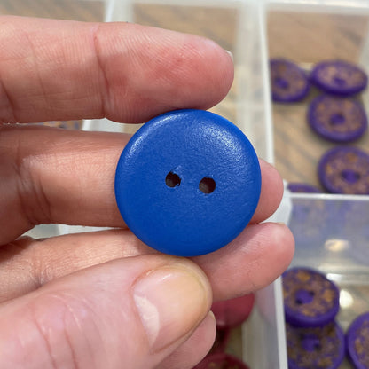 Hand holding a blue button with a background of button storage container.