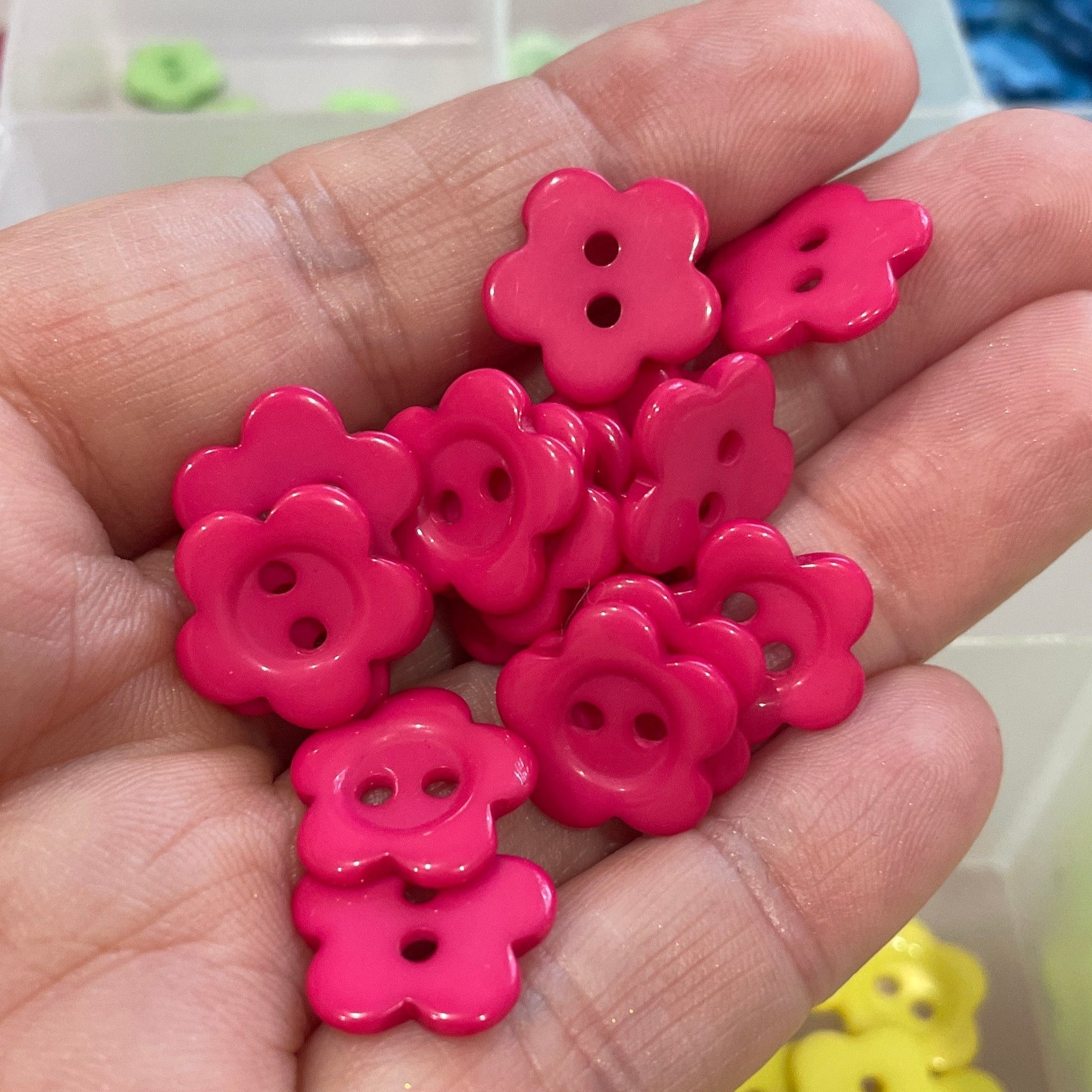 Hand holding pink flower-shaped buttons with a container of colorful buttons in the background.