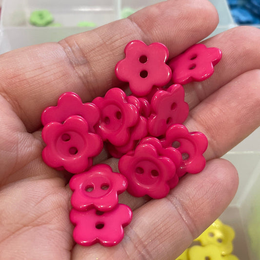 Hand holding pink flower-shaped buttons with a container of colorful buttons in the background.
