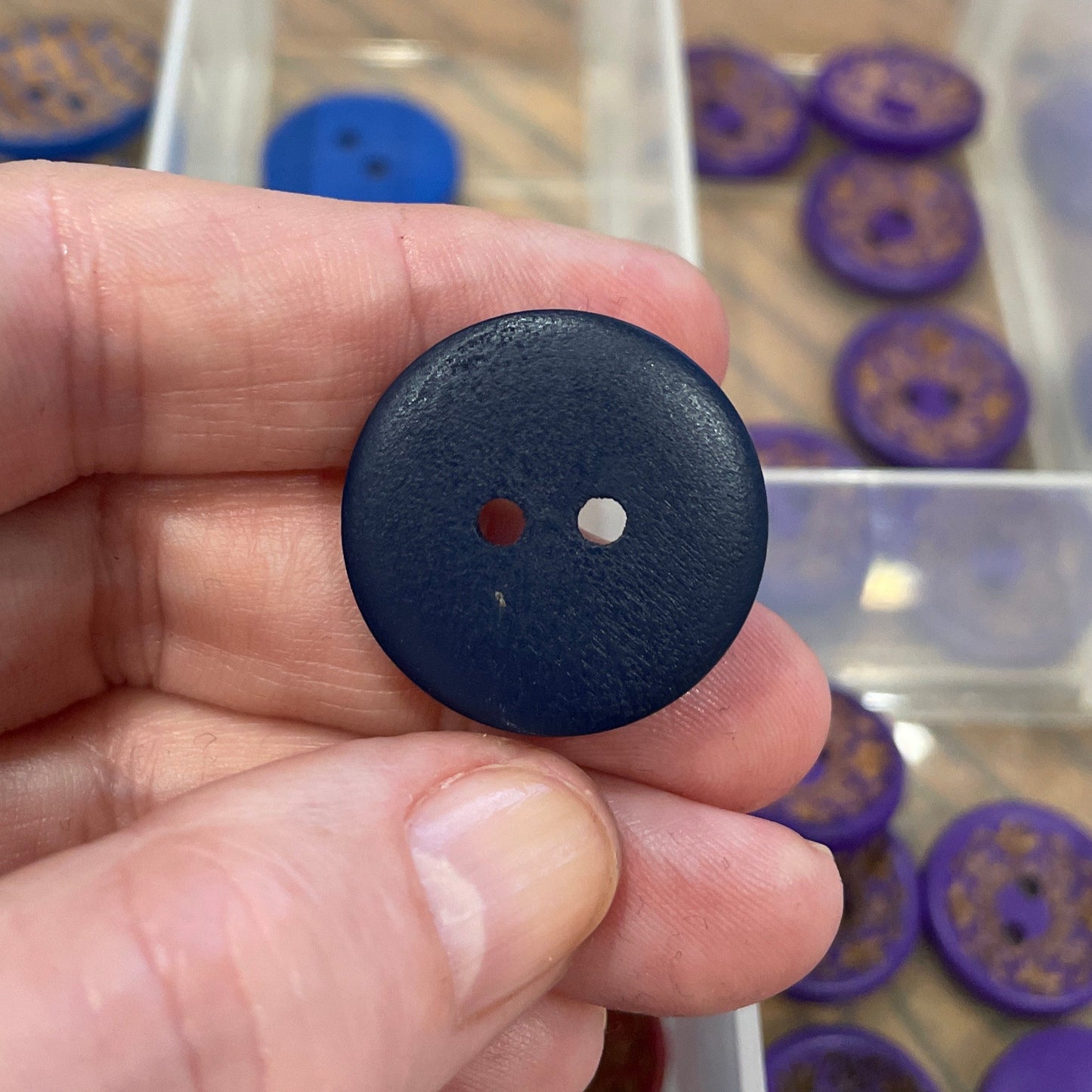 Hand holding a blue button with a white center, against a background of organized buttons in a clear container.