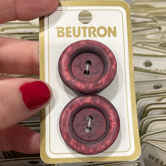Two maroon buttons on a Beutron card held by a hand with red nail polish.