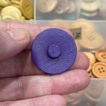 Hand holding a purple button with a container of assorted buttons in the background
