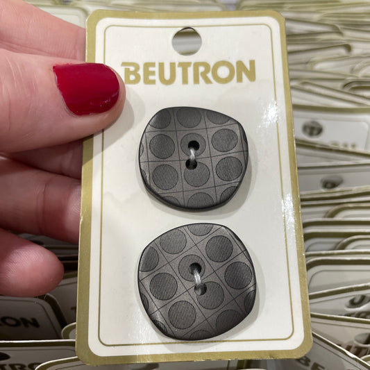 Two gray buttons on a Beutron branded card held by a hand with red nail polish.