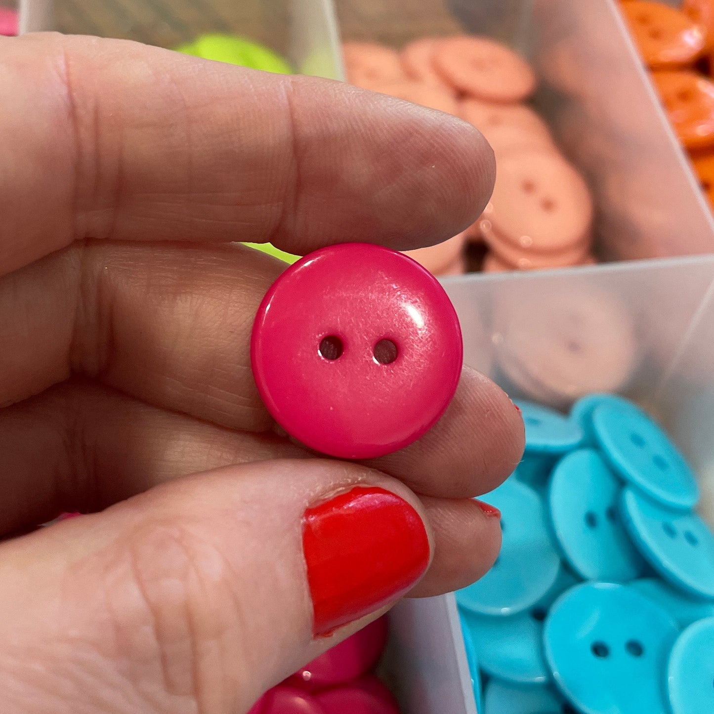Hand holding a pink button with containers of buttons in the background