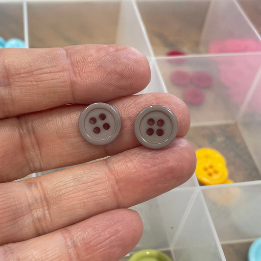 Hand holding two gray buttons with a clear container of colorful buttons in the background.