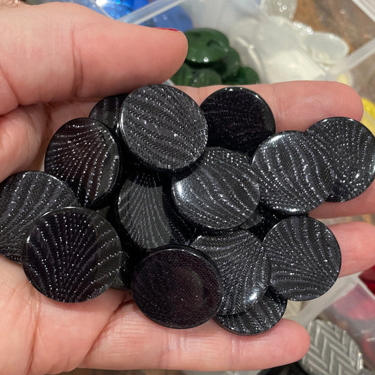 Hand holding black rhinestone buttons with a container of various colored rhinestones in the background.