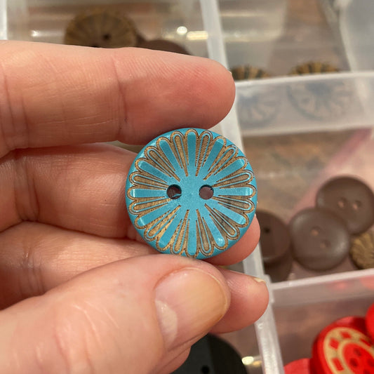 Hand holding a blue decorative button with a container of assorted buttons in the background.