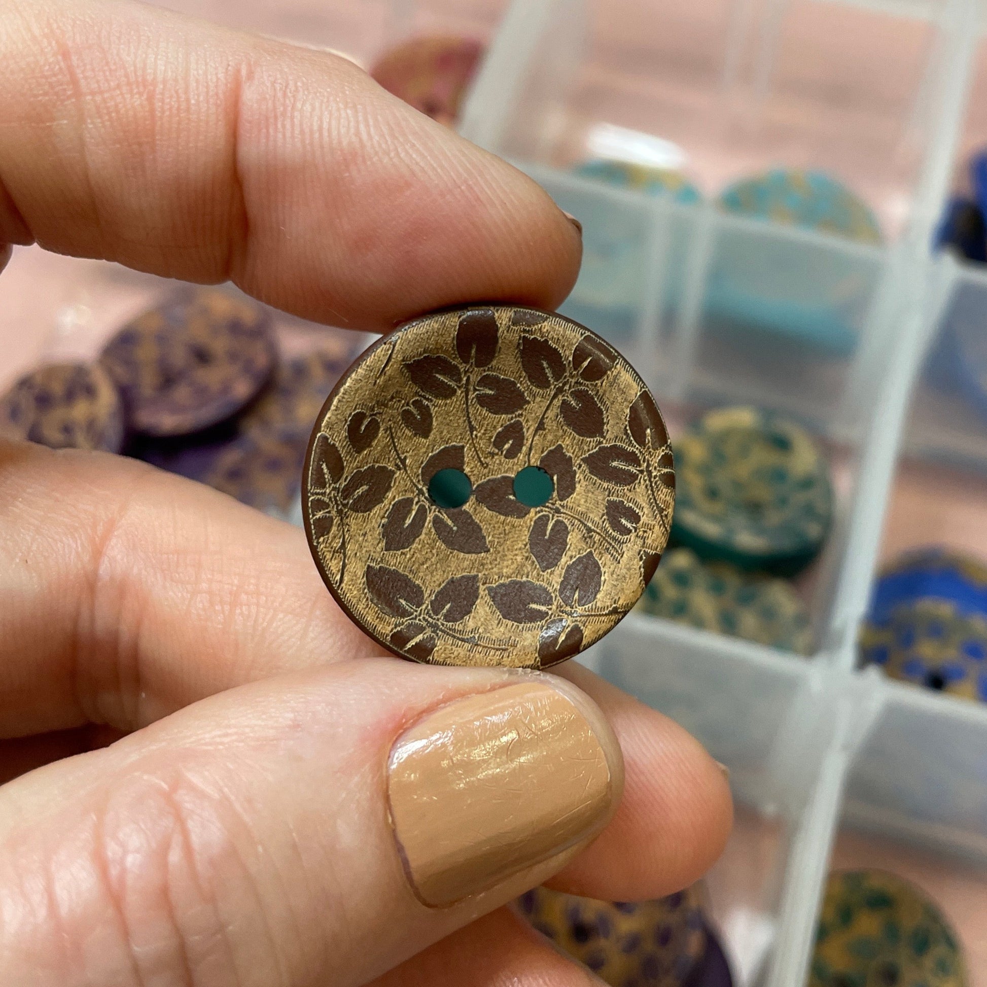 Hand holding a decorative button with intricate design, in front of a container filled with various buttons.