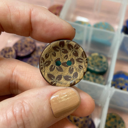 Hand holding a decorative button with intricate design, in front of a container filled with various buttons.