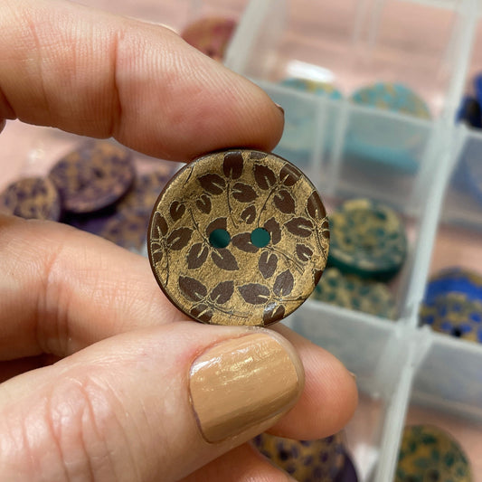 Hand holding a decorative button with intricate design, in front of a container filled with various buttons.
