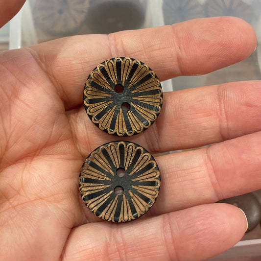 Hand holding two bronze floral-patterned buttons with a container of assorted buttons in the background.