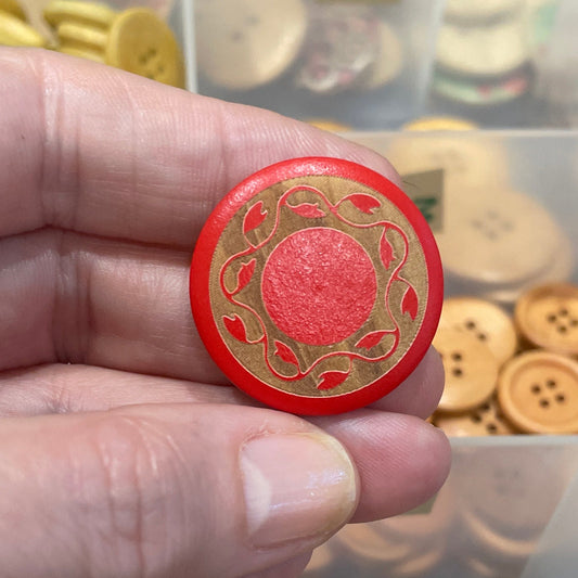 Hand holding a red button with gold design in front of a container of various buttons.