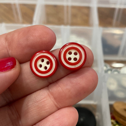 Two red buttons with white centers held between fingers in front of a container of buttons.
