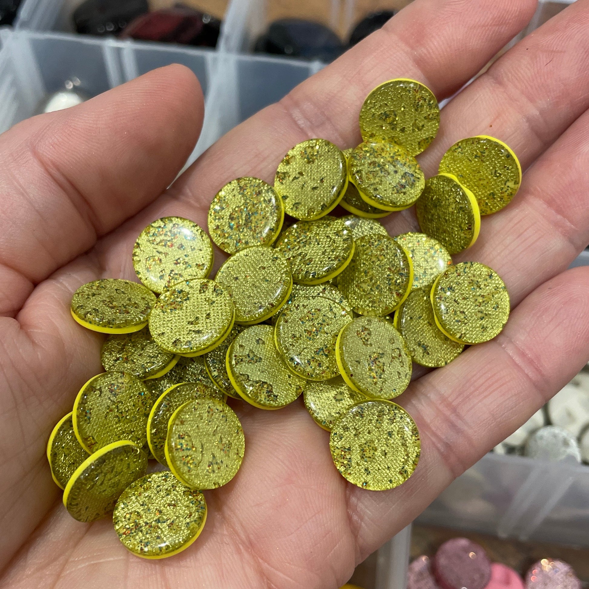 Hand holding small round green glittery objects with a container of similar objects in the background.