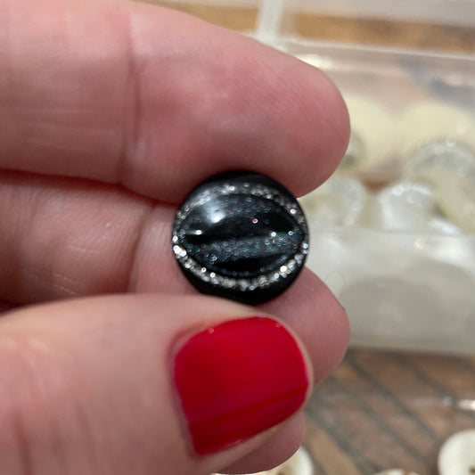 Black button held by a hand with red nail polish, with a container of buttons in the background.