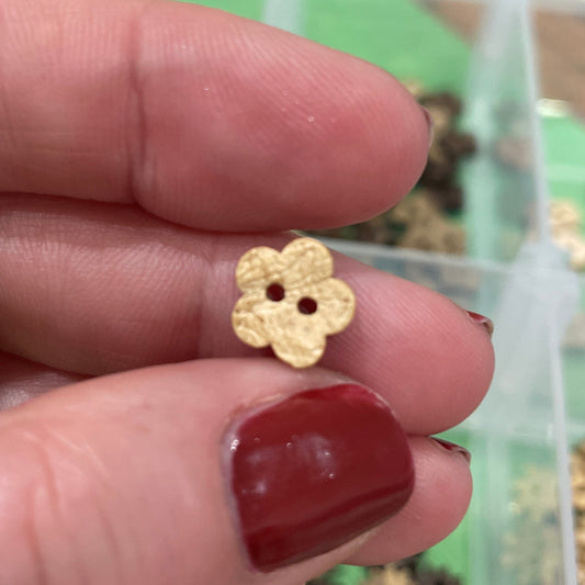 Gold flower-shaped button  held between fingers with a container of similar buttons in the background.