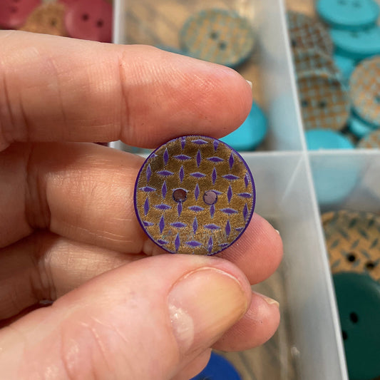 Hand holding a round button with a unique pattern, with a container of various buttons in the background.