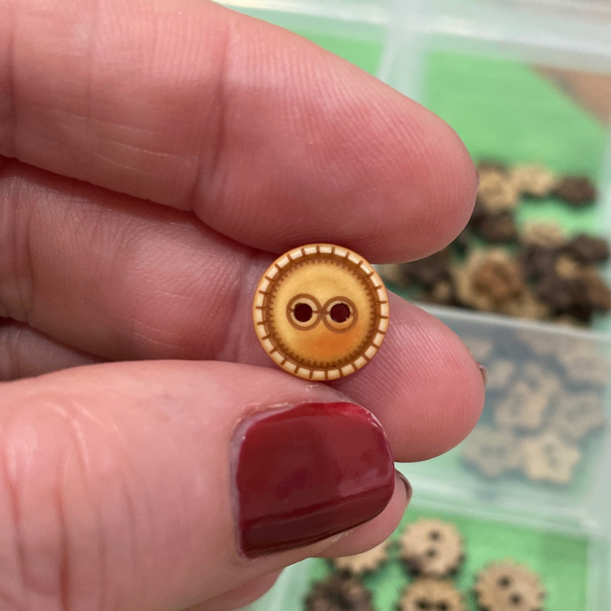 Gold button held by a hand with red nail polish, against a background of more buttons in a container.