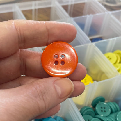 Hand holding an orange button with a background of colorful buttons in containers.