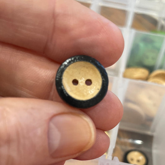 Hand holding a black and gold button with a container of colorful buttons in the background