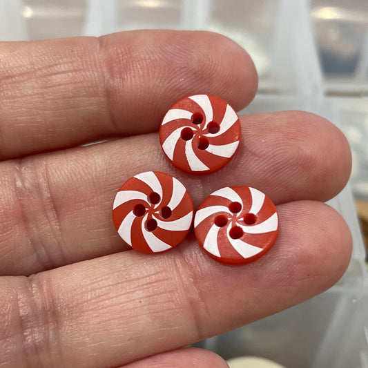 Three red and white button earrings held between fingers with a container of buttons in the background.