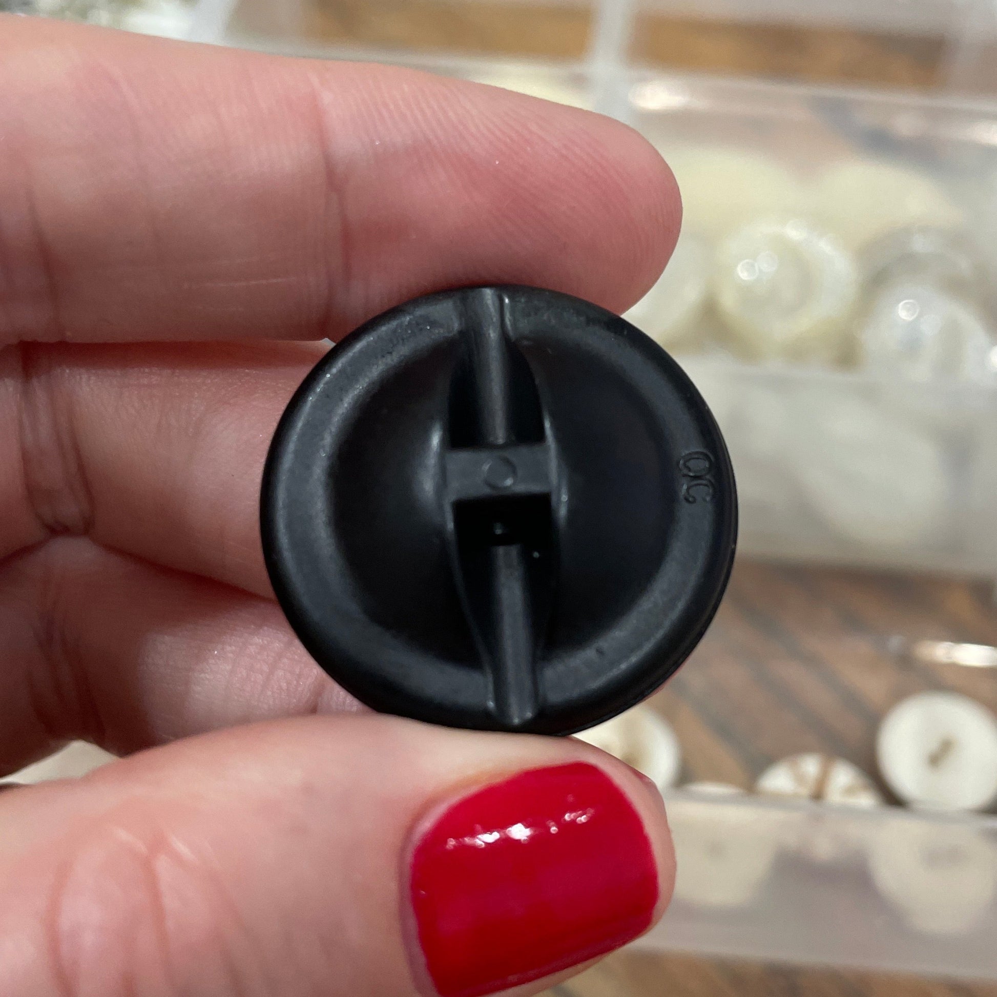 Black plastic component held by a hand with red nail polish, with a clear container of small items in the background.