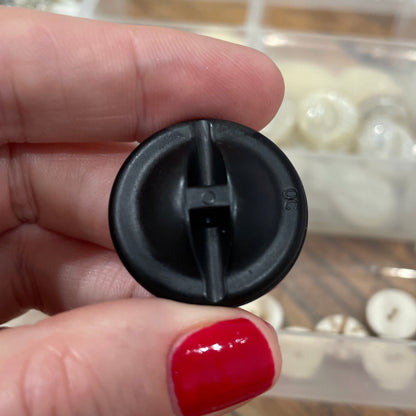 Black plastic component held by a hand with red nail polish, with a clear container of small items in the background.