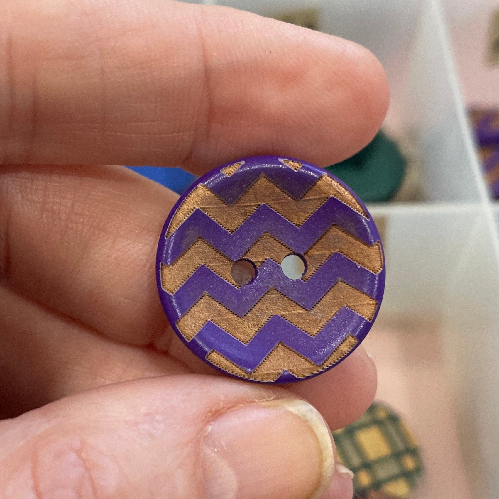 Hand holding a purple and gold zigzag patterned button with a blurred background of a button collection.