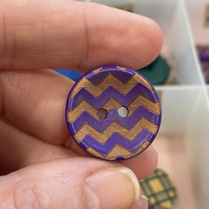 Hand holding a purple and gold zigzag patterned button with a blurred background of a button collection.