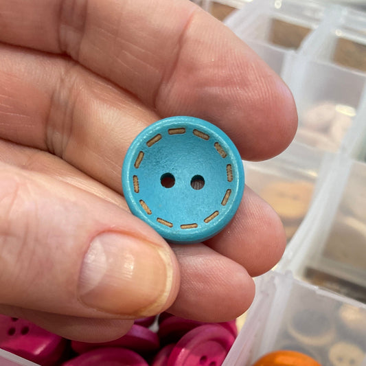 Hand holding a blue button with a container of colorful buttons on a wooden surface