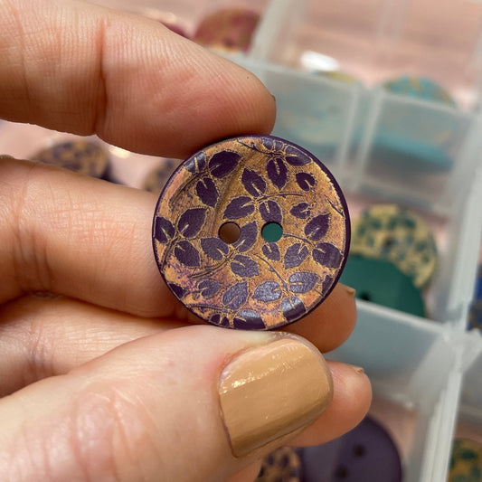 Hand holding a decorative button with a floral pattern, against a background of a clear container with compartments.