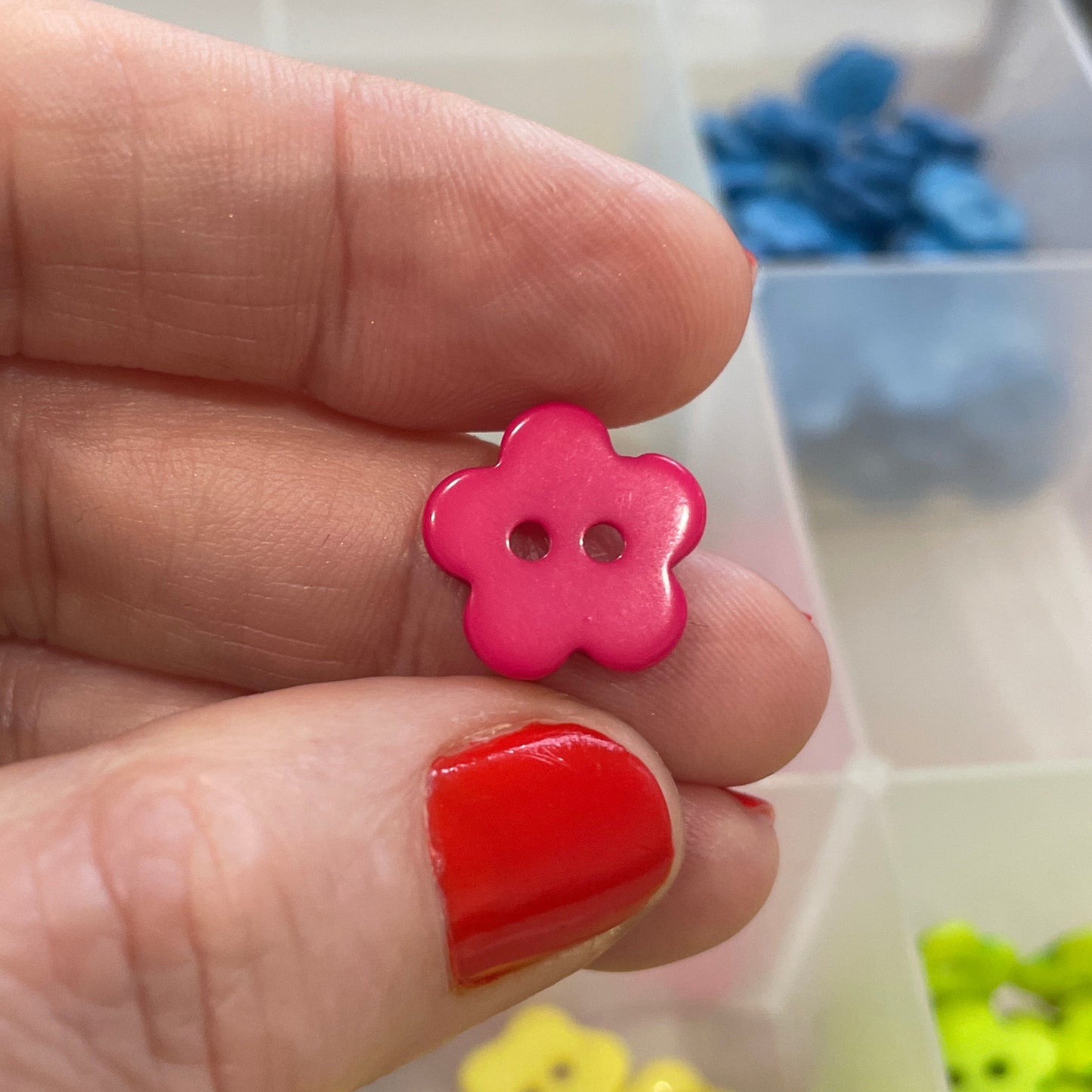 Hand holding a pink flower-shaped button with a background of colorful buttons in a container.
