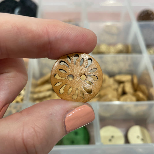 Hand holding a decorative wooden button with intricate cut-out design, against a background of similar buttons in a container.