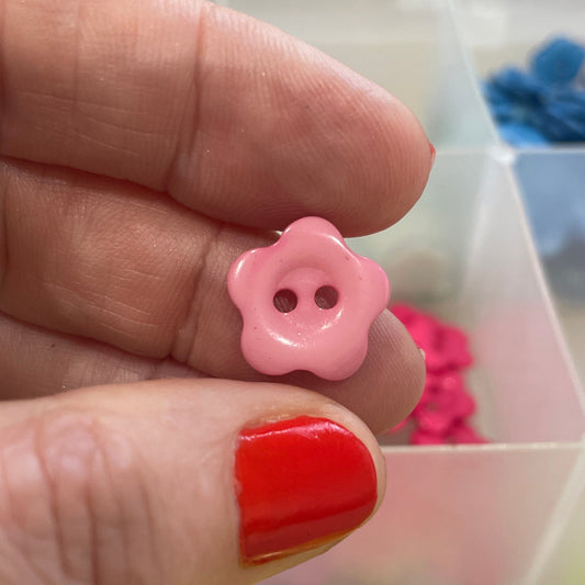 Hand holding a pink flower-shaped button with a clear container of colorful buttons in the background.