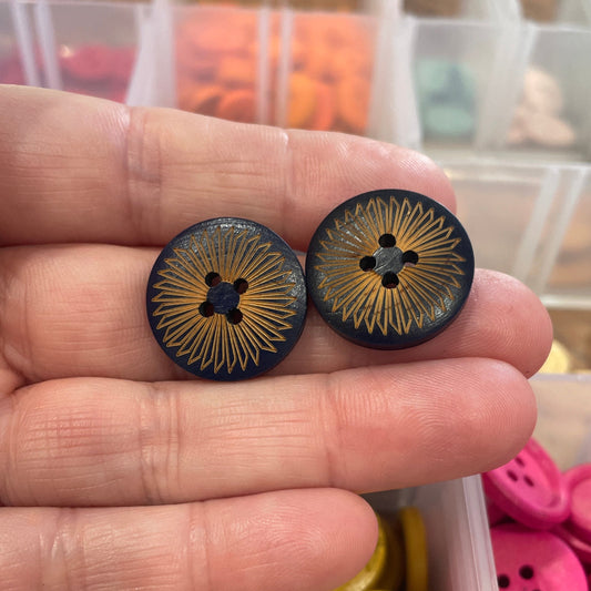 Two black buttons with gold patterns held between fingers, with a container of colorful buttons in the background.