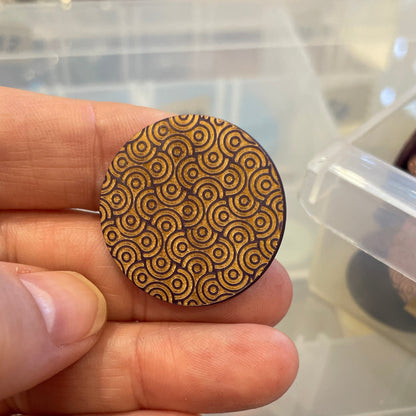 Hand holding a wooden button with a pattern, surrounded by other buttons in a clear container.