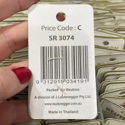 Hand holding a product tag with a barcode and text, with shelves of similar products in the background.