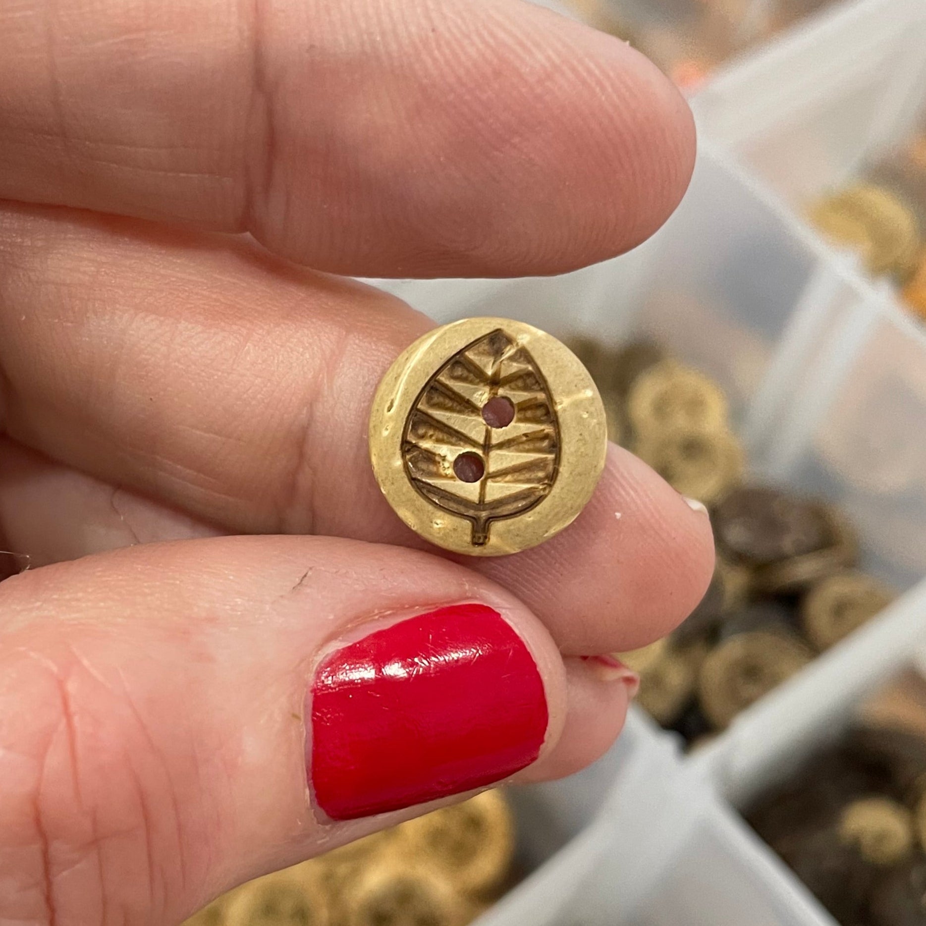Hand holding a button with a detailed design, against a background of various buttons in a container.