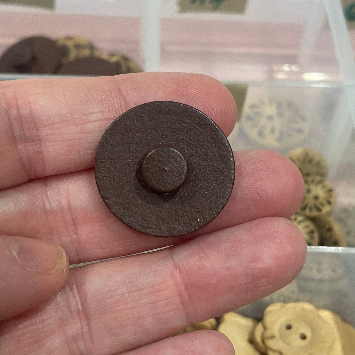 Hand holding a small button with a background of button assortment in a box.