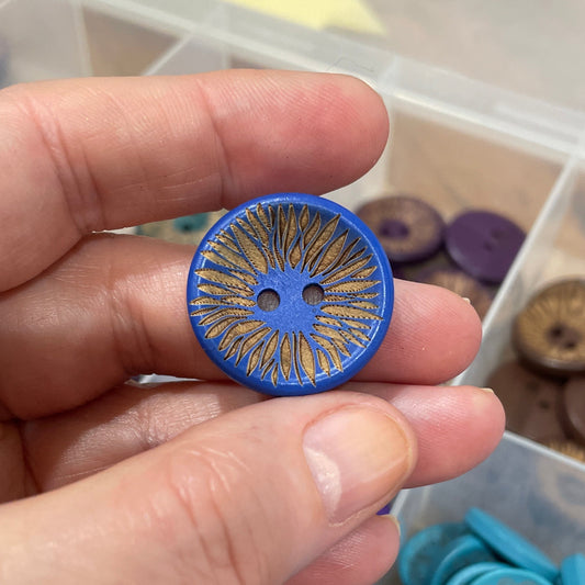 Hand holding a blue button with intricate design, with a container of buttons in the background.