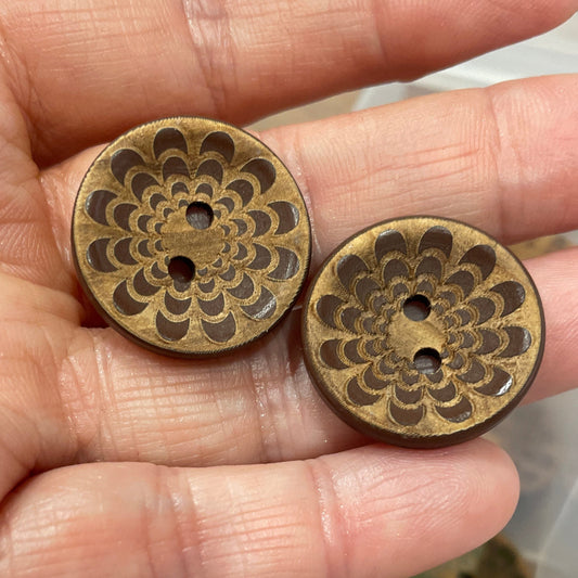 Bronze buttons with intricate designs held in a hand, with a container of similar buttons in the background.