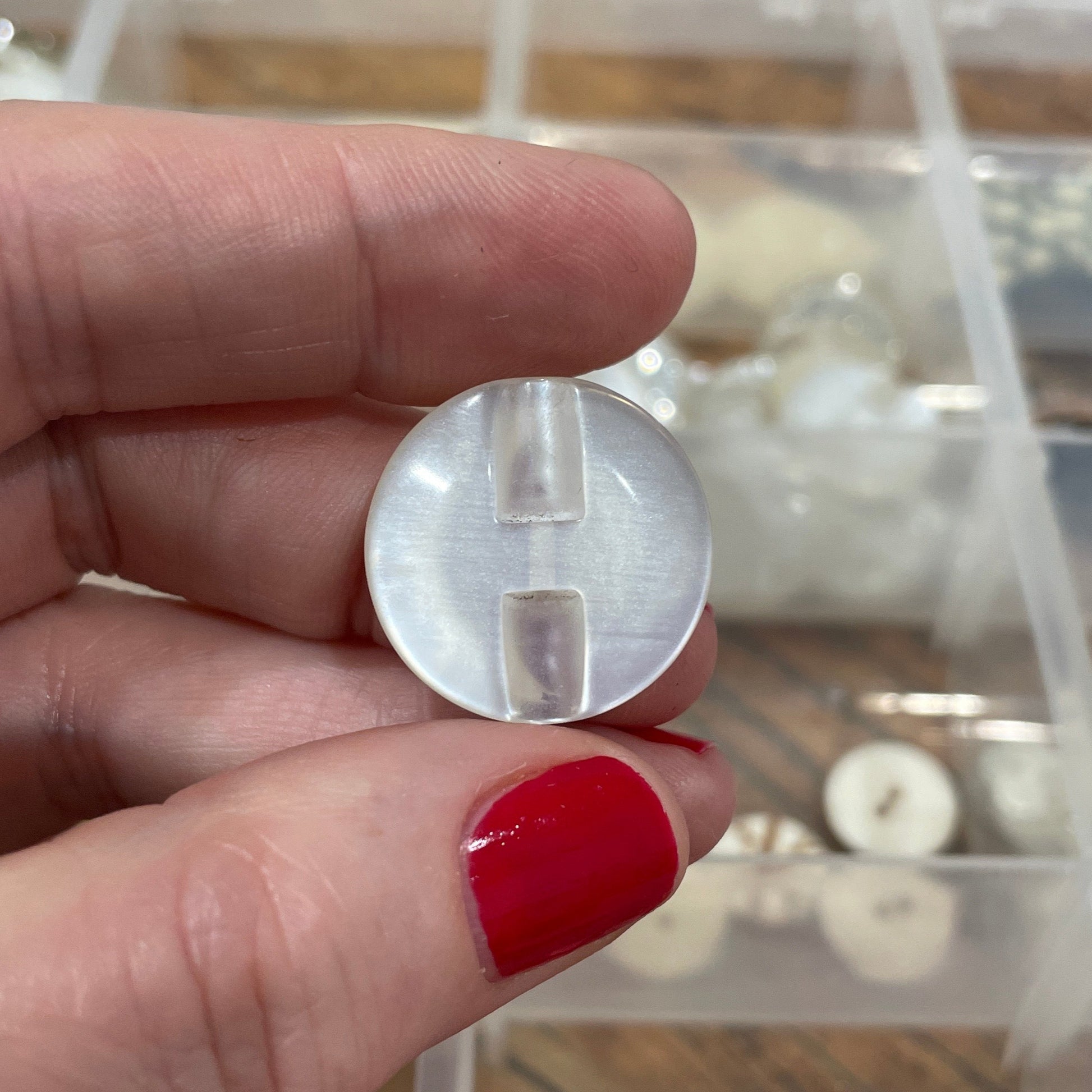 Hand holding a clear plastic component with a container of buttons in the background