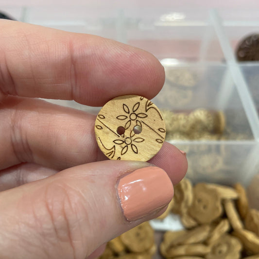 Hand holding a gold button with floral design in front of a container of assorted buttons.