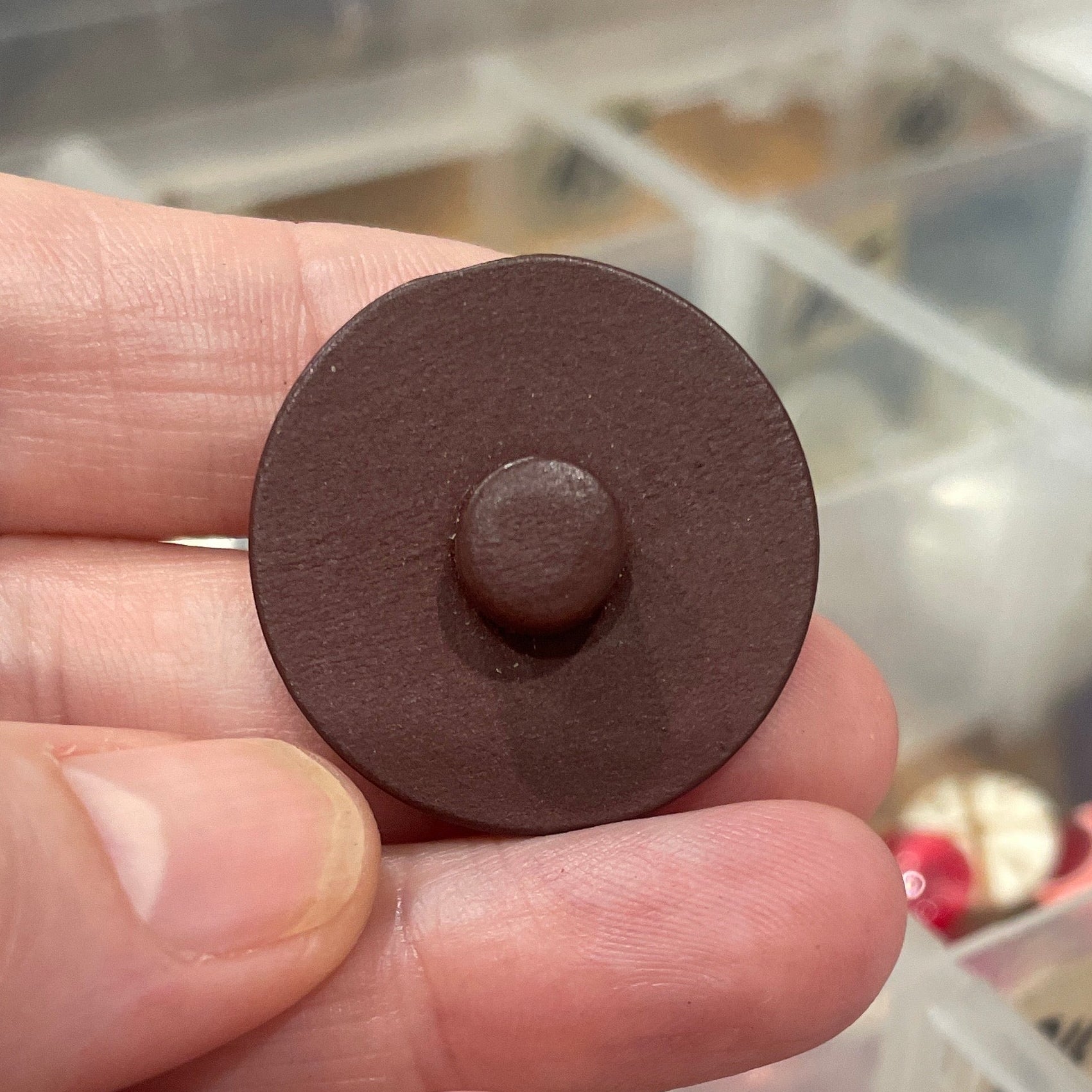 Brown buttons held between fingers with a blurred background of containers.
