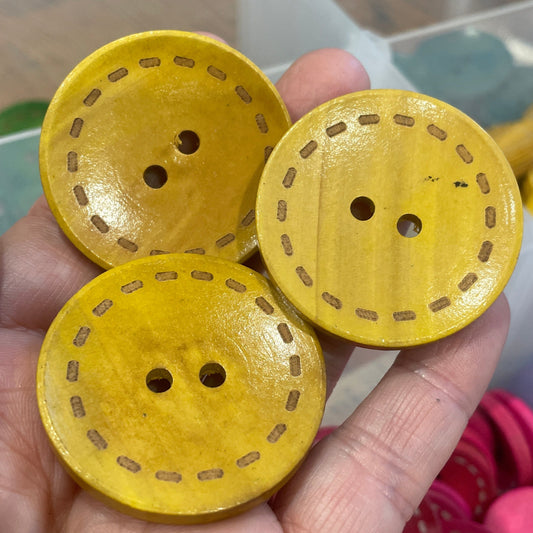 Yellow buttons with holes held by a hand, with a container of colorful buttons in the background.