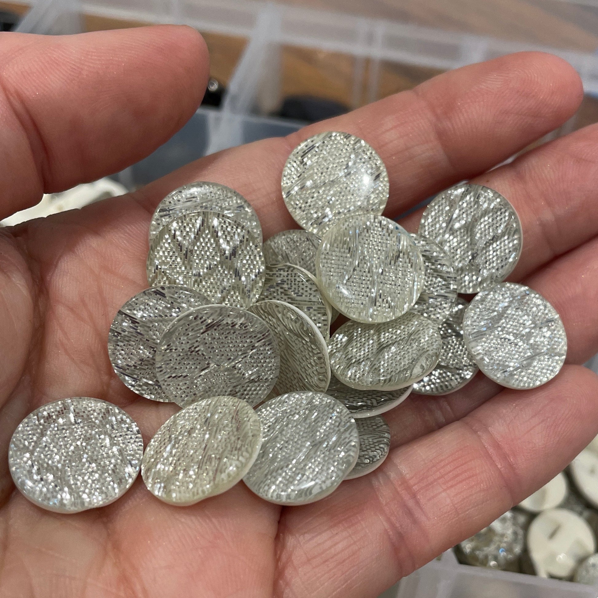 Hand holding silver oval beads with a container of colorful buttons in the background