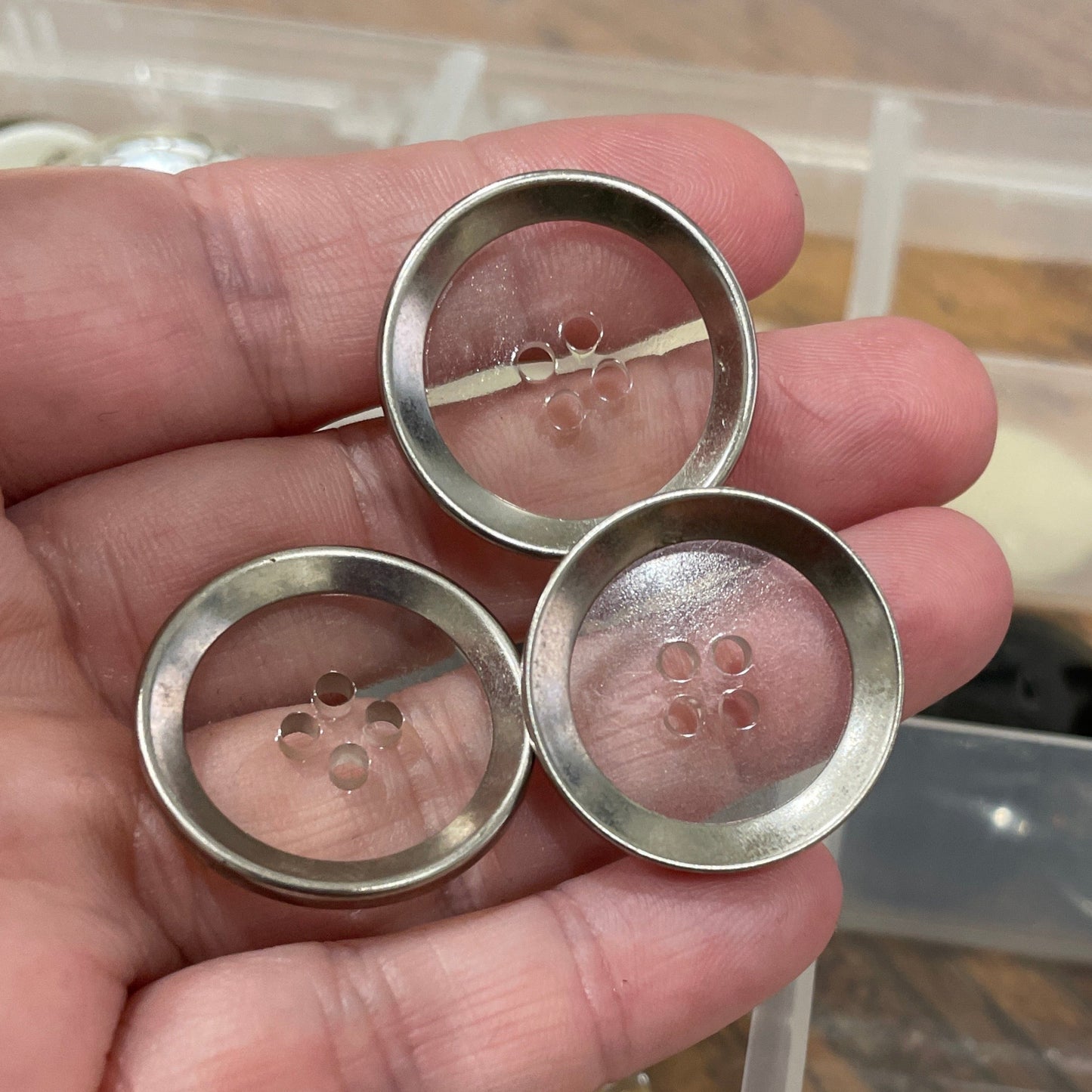 Hand holding three silver buttons with a clear container of buttons in the background.