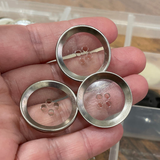 Hand holding three silver buttons with a clear container of buttons in the background.