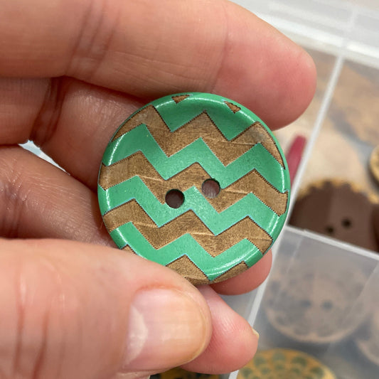 Hand holding a green and brown zigzag patterned button with a container of similar buttons in the background.