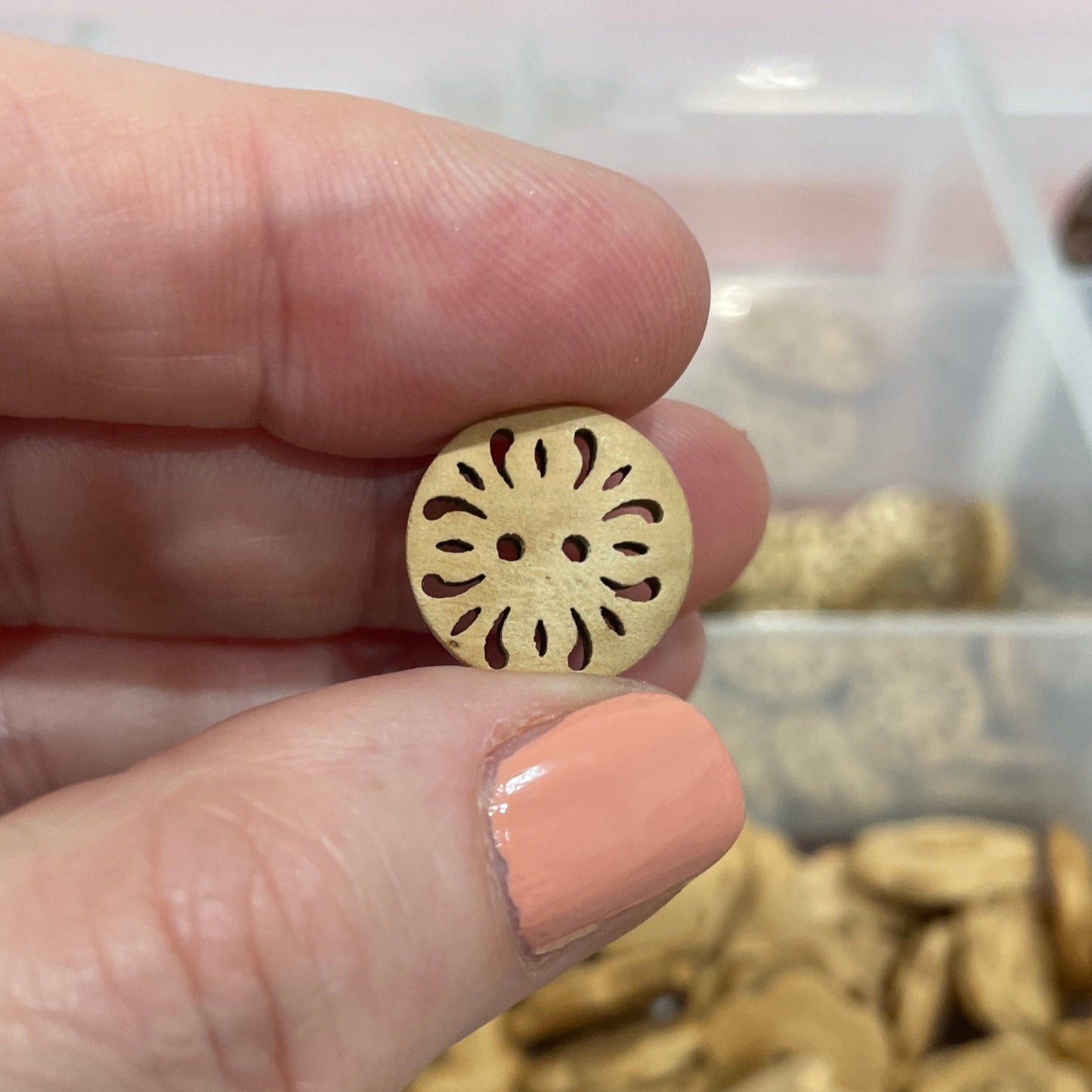 Hand holding a gold button with intricate design, with a container of buttons in the background.